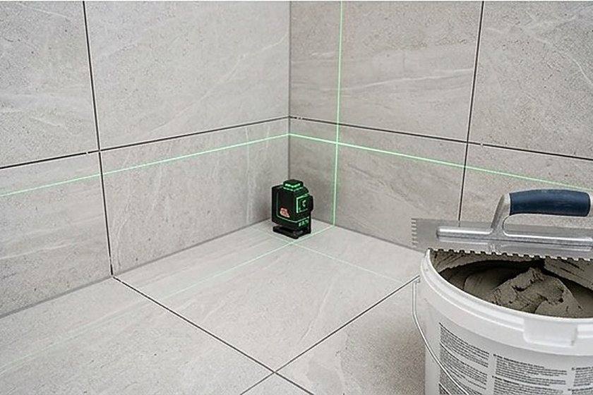 Großformatige Wandfliesen, Kreuzlinienlaser und Zahnkelle auf dem Fliesenkleber-Eimer